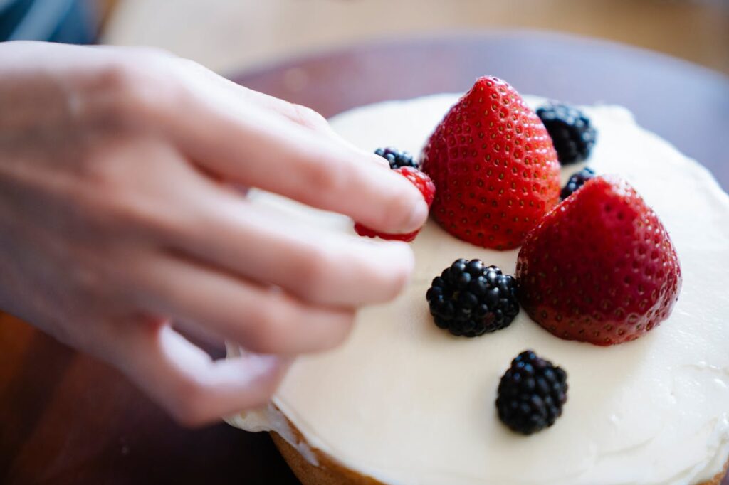 Hand placing berries on a cake with fresh frosting, highlighting baking skills.